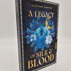 Der Text "A Legacy of Silk & Blood" von Stefanie Santer ziert das Cover. Eine Illustration zeigt blaue Blumen und einen Schmetterling.
