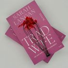 Zwei rosa Buchcover, darauf "SARAH LANGAN", "TRAD WIFE", "SIE HAT NUR AUF DICH GEWARTET". Ein Schneebesen sowie rote Farbe sind darauf platziert.