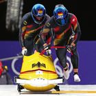 Zwei Bobfahrer mit "GER" Helmen, schwarzer Kleidung mit Deutschlandflagge, stoßen gelben Bob an, ein Adler-Logo ist zu sehen.
