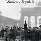 "Wankende Republik" in großer Schrift über einer Menschenmenge, die in historischer Kleidung marschiert.