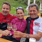 Drei Menschen in Sportkleidung mit "Ischgl Ironbike"-Text, lächelnd beim Anstoßen mit Weingläsern in einer grünen Umgebung.