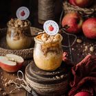 "Apfelkuchen im Glas" steht auf einem runden Schild. Zwei Gläser mit Apfelkuchen, umgeben von Äpfeln und rustikalen Dekoelementen.
