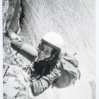 Eine Person klettert lächelnd an einer Felswand, trägt einen Schutzhelm und einen Rucksack, wirkt abenteuerlustig.