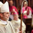 Ein Bischof in festlicher Kleidung, trägt einen Hirtenstab und eine Mitra, umgeben von anderen Geistlichen in einer Zeremonie.