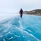 Ein Mensch in bunter Winterkleidung geht auf einer spiegelglatten, blauen Eisfläche, umgeben von verschneiter Landschaft.