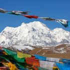 Verschneite Berge unter klarem Himmel. Bunte Gebetsfahnen wehen im Vordergrund.