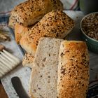 Mehrkornbrötchen auf Tuch mit Kruste aus Sesam und Körnern, daneben ein Messer und eine Schüssel mit Körnern.