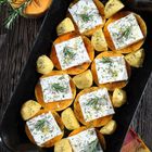 Kürbisscheiben und Kartoffeln, belegt mit Fetastreifen und Rosmarin, in einer schwarzen Auflaufform mit roten Griffen.