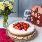 Ein Kuchen mit Erdbeeren und Puderzucker, daneben gelbe Blumen und eine rote Keramikhaus-Deko auf einem Tisch.