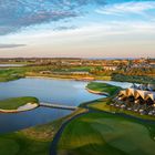 Ein Golfplatz mit Wasserhindernissen, grüner Landschaft, modernen, zeltartigen Gebäuden und einem klaren Abendhimmel.