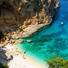 Keine Texte sichtbar. Türkisblaues Meer, kleine sandige Bucht, umgeben von Felsen und grünen Bäumen. Ein Boot im Wasser.