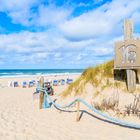 Schild mit der Zahl 16 auf einer Düne; dahinter Strandkörbe, Sand, Meer und blauer Himmel mit Wolken.