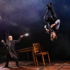 Zwei Personen in einem dramatischen Duell, eine steht aufrecht mit leuchtendem Stab, die andere schwebt kopfüber in der Luft.