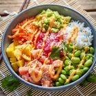 Eine Schüssel mit Reis, Lachs, Mango, Edamame, Karotten, Avocado, Sesam und Soße auf einer Bambusmatte.
