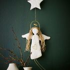 Ein Engel aus weißem Material mit goldener Heiligenschein hängt unter einem Stern vor einem grünen Hintergrund; unten weiße Vasen.