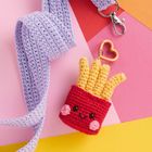 Häkel-Anhänger in Form von Pommes in roter Tüte mit lachendem Gesicht, auf buntem Hintergrund mit lila Band und Karabiner.