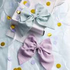 Zwei große Stoffschleifen liegen auf Seidenpapier; eine in Pastellblau, die andere in Flieder. Umgeben von buntem Konfetti.