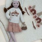 Eine gestrickte Puppe mit Zöpfen trägt einen cremefarbenen Pullover, einen rosa Rock und hält eine kleine Tasche. Daneben gestrickte Blumen.
