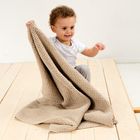 Ein Kind mit lockigem Haar und hellem T-Shirt spielt auf einem Holzboden mit einer großen, beigen Strickdecke.