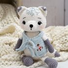 Gehäkeltes Kuscheltier als Bär, hellblaue Weste mit Schneemannmotiv, auf cremefarbener Decke sitzend.