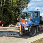 Blauer Lastwagen mit großer Bürste vorne, roten Warnflaggen und grünem Wald im Hintergrund.