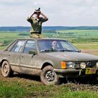 Eine Person sitzt auf einem alten, schlammigen Auto und schaut durch ein Fernglas über weites Feld.
