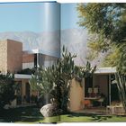 Ein modernes Haus mit Flachdach und Steinkamin, umgeben von Kakteen und Bäumen, mit Bergen im Hintergrund.
