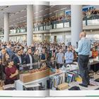 "Roots 45" rechts unten. Ein Mann spricht mit Mikrofon vor einer großen Menschenmenge in einem modernen Büro.