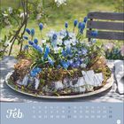 Februar-Kalender mit Frühlingsblumen auf einem Holztisch im Garten, umgeben von blühenden Zweigen und einer Holzbank.