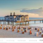 FEBRUAR. Strandkörbe auf Sand vor einer Seebrücke mit einem Gebäude, dahinter das Meer und eine Wolkenformation.