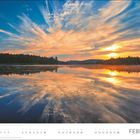FEBRUAR. Ein ruhiger See mit spiegelnder Wasseroberfläche, umgeben von Wald, Sonnenuntergang.
