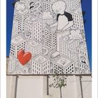 02; Illustration eines riesigen Kindes, das über eine Stadt schaut, mit Gebäuden, Wolken und einem roten Herz an der Wand.