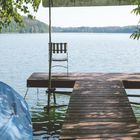 Eine hölzerne Anlegestelle am See mit einem einsamen Stuhl. Blätterrahmen im Vordergrund.