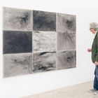 Mann betrachtet Kunstwerk aus neun verschiedenen grauen Quadraten an einer weißen Wand.