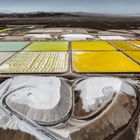 Salzpfannen mit intensiven Gelb- und Grüntönen, daneben weiße und braune Erdhügel, umgeben von einer weitläufigen Wüstenlandschaft.