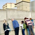 Text: "Reconstruction of the feared 'Death Strip' at the Berlin Wall Memorial." Personen blicken durch eine Mauer.