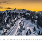 Februar 2024, ein verschneiter Bergweg schlängelt sich durch mit Bäumen gesäumte Hügel bei Sonnenuntergang.