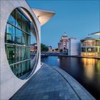 Text fehlt. Moderne Architektur mit großen runden Fenstern, Wasser und Fahnen bei Dämmerung, im Hintergrund ein historisches Gebäude.