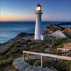 Ein Leuchtturm beleuchtet die Dämmerung an einer felsigen Küste, umgeben von einem Holzweg und Pflanzen.