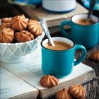 Geöffnete Bücher, zwei blaue Tassen mit Kaffee, ein Schälchen Gebäck, Löffel in Tassen, Holztisch.