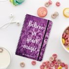 „Believe in your magic“, Notizbuch in Violett mit floralen Mustern, umgeben von buntem Frühstücksgetreide und Ohrstöpseln.