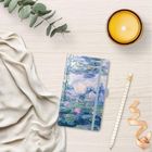 Notizbuch mit Seerosenmotiv, daneben eine brennende Kerze, ein Bleistift und ein Stofftuch auf hellem Holz.