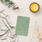 Grünes Notizbuch mit floralen Mustern und "2024" Prägung, daneben eine Kerze, ein Bleistift, Eukalyptus und ein beiges Tuch.