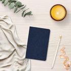 Ein dunkelblaues Notizbuch mit floralem Muster liegt auf hellem Holz, daneben eine Kerze, ein Bleistift, Band, Tuch und Eukalyptus.