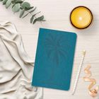 Notizbuch mit eingeprägter Palme und Text "2024". Daneben liegen ein Bleistift, Eukalyptuszweig, Kerze und Stoff.