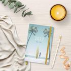 2027 Mein Jahr. Ein Notizbuch mit Palmen-Illustration, gelbes Band, neben Kerze, Stift, Schleife und Stoff.