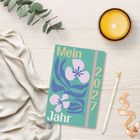„Mein Jahr 2027“ steht auf einem Kalender mit Blumenmotiv auf hellem Tisch, neben Kerze, Bleistift und Stoff.
