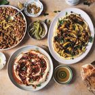 Verschiedene Teller mit Hummus, Bohnen, gegrilltem Gemüse, Brot, Gewürzen und Zitronenscheiben auf einem hellen Untergrund.
