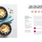 "Soja-Chia-Müsli mit Apfel und Hüttenkäse". Zwei Schalen mit Müsli, Apfelspalten, auf grünem Tuch, daneben Kardamomschoten.