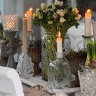 Mehrere brennende Kerzen in Kristallhaltern umgeben eine Vase mit weißen Blumen auf einem Esstisch.